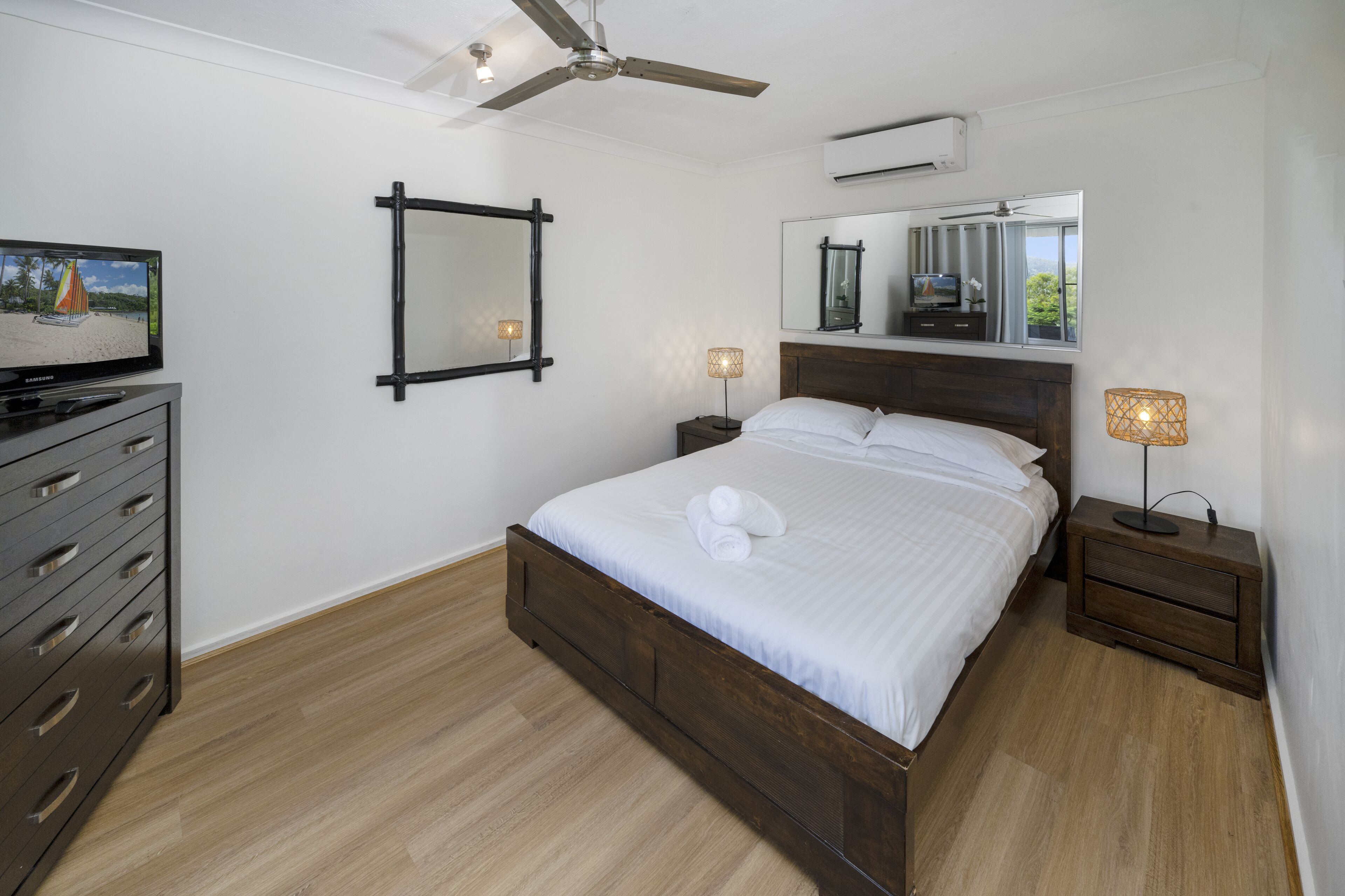 Ein Schlafzimmer mit Bett, Kommode, Fernseher, Deckenventilator, Spiegel und Nachttisch mit Lampe.