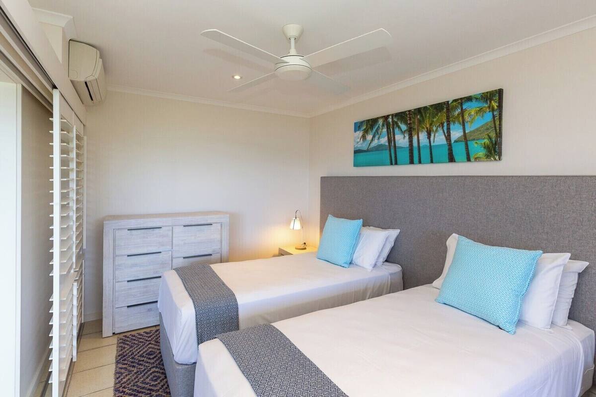 A hotel room with two beds, a ceiling fan, a painting of a beach, and a white wardrobe.