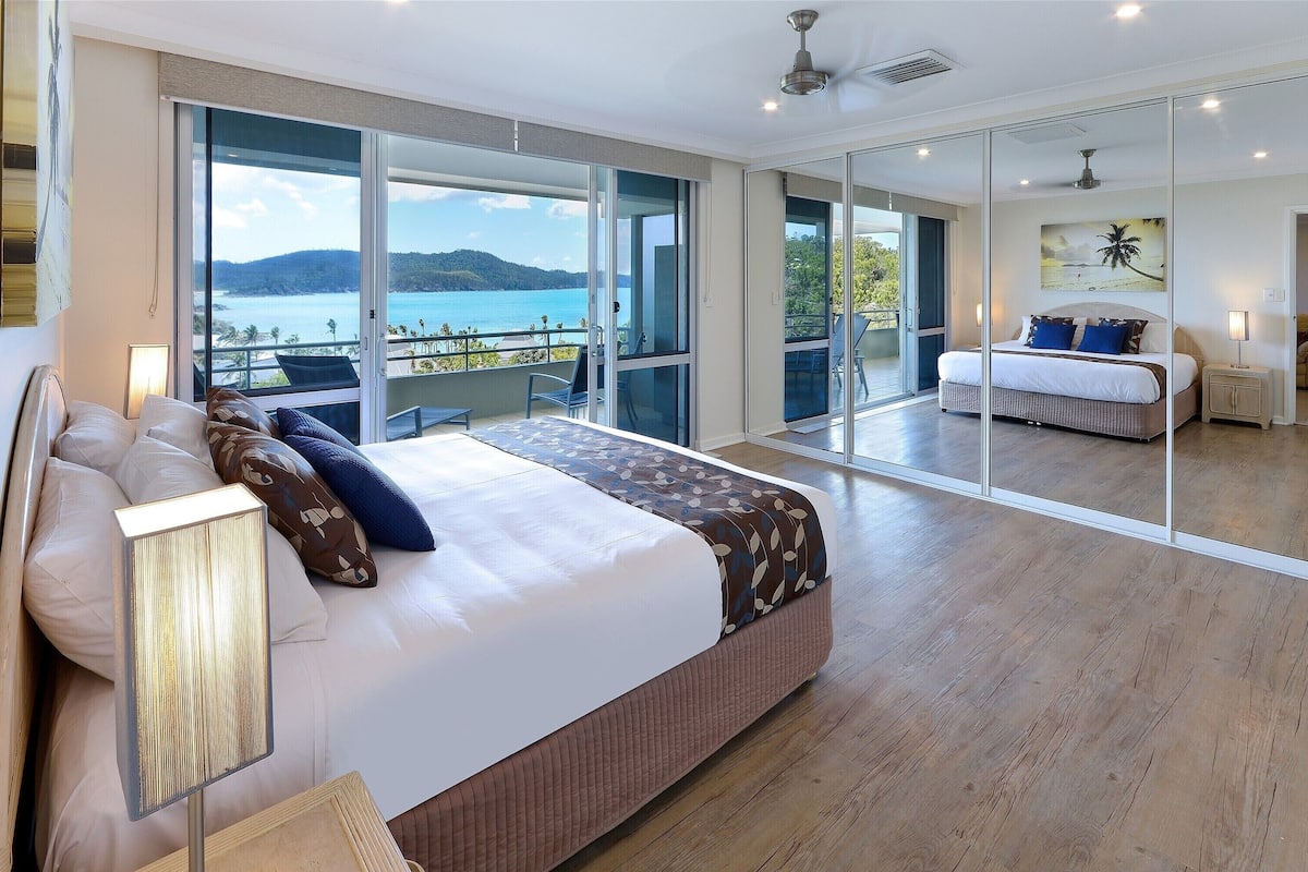 A bedroom with a large bed, a nightstand, a lamp, and a view of the sea through large windows.