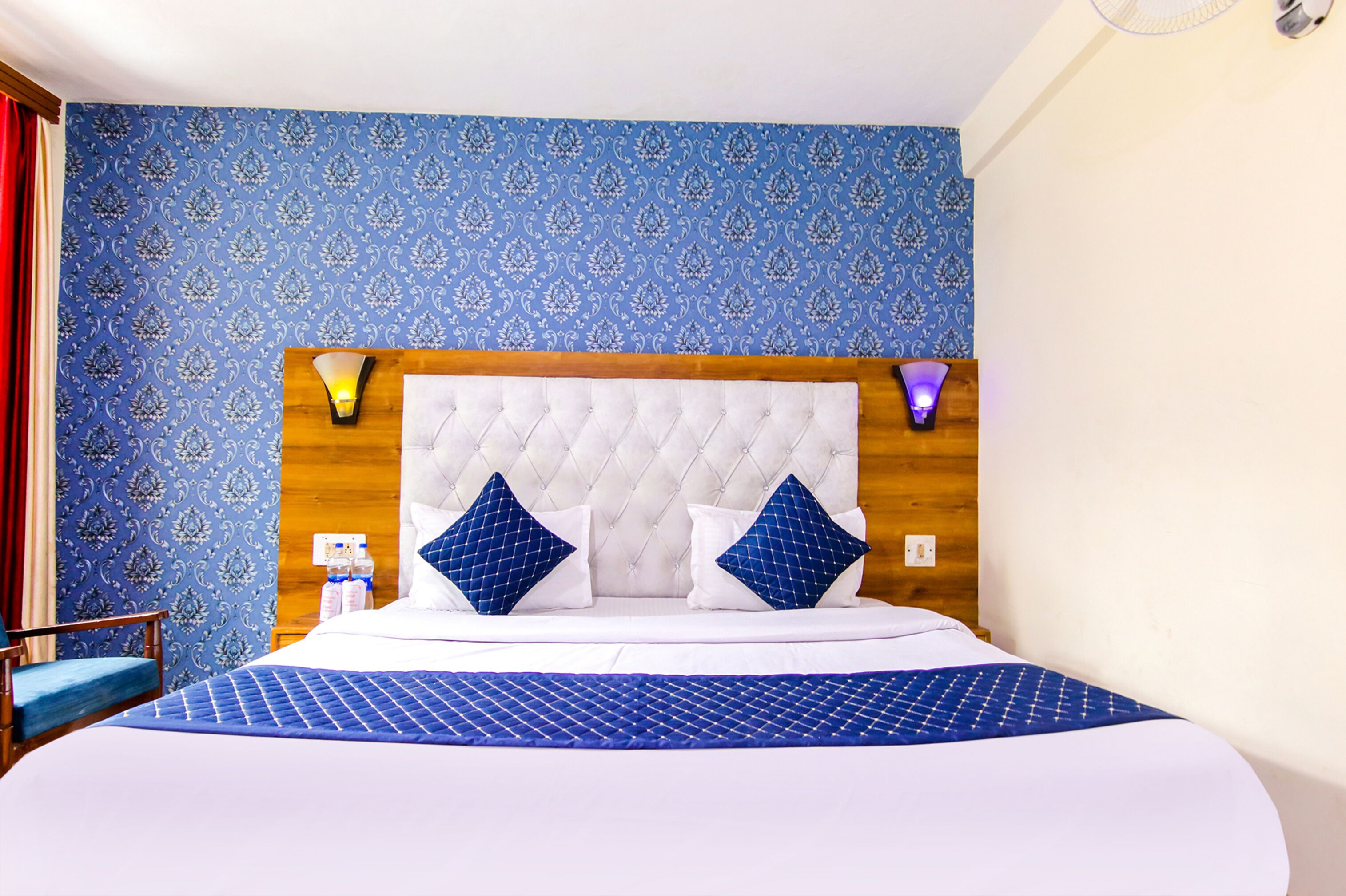 A hotel room with a large bed, blue patterned wallpaper, a wooden headboard, two bedside lamps, and a blue bedspread.