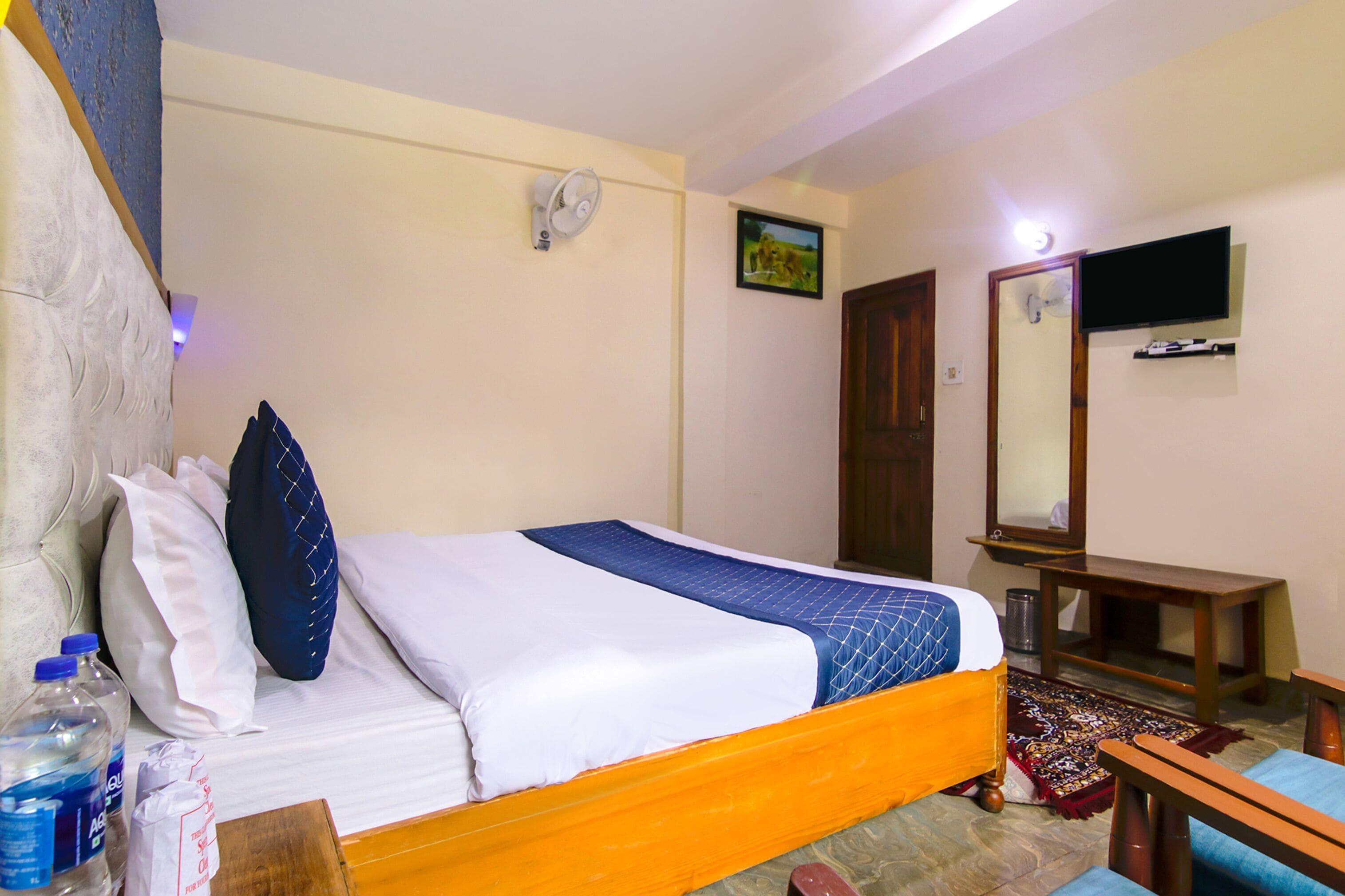 A hotel room with a bed, pillows, a bedside table, a mirror, a television, and a small wooden table.