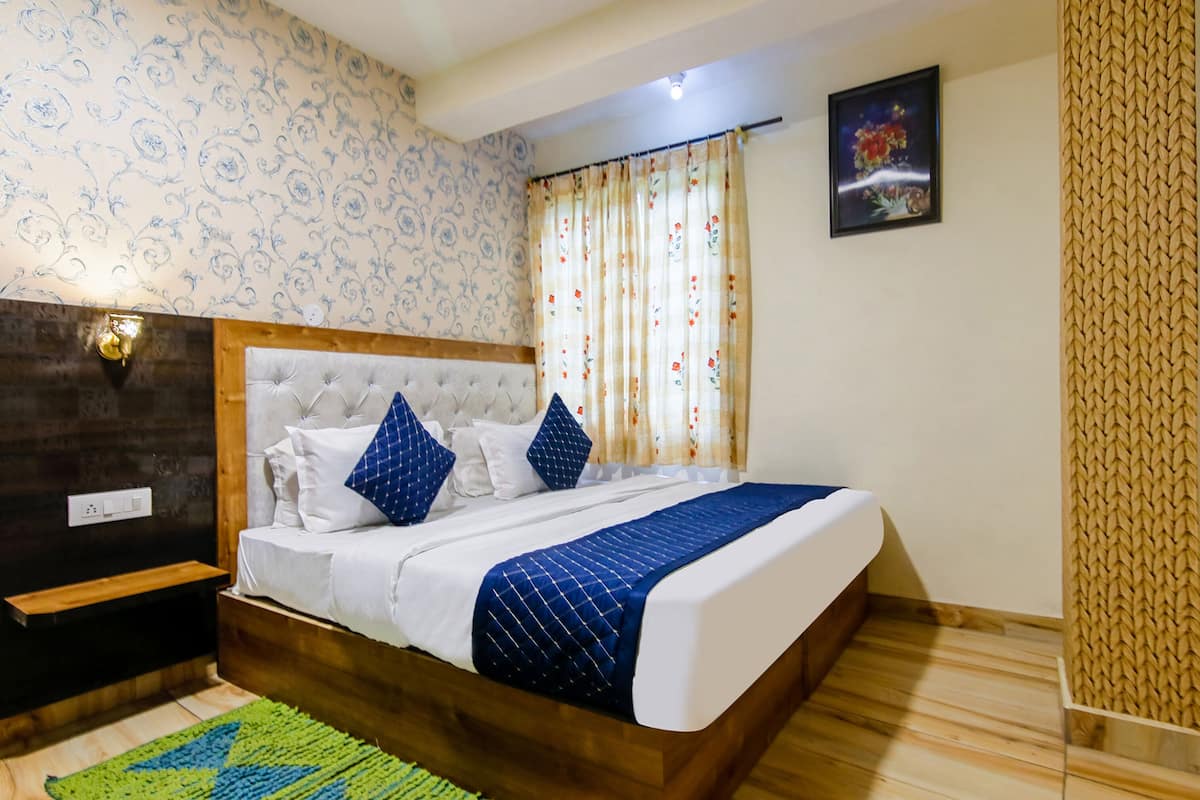 Ein ordentlich eingerichtetes Schlafzimmer mit einem Holzkopfteil, einem Bett mit weißblauer Bettwäsche, einem grünen Teppich und einem Vorhang mit Blumenmuster.