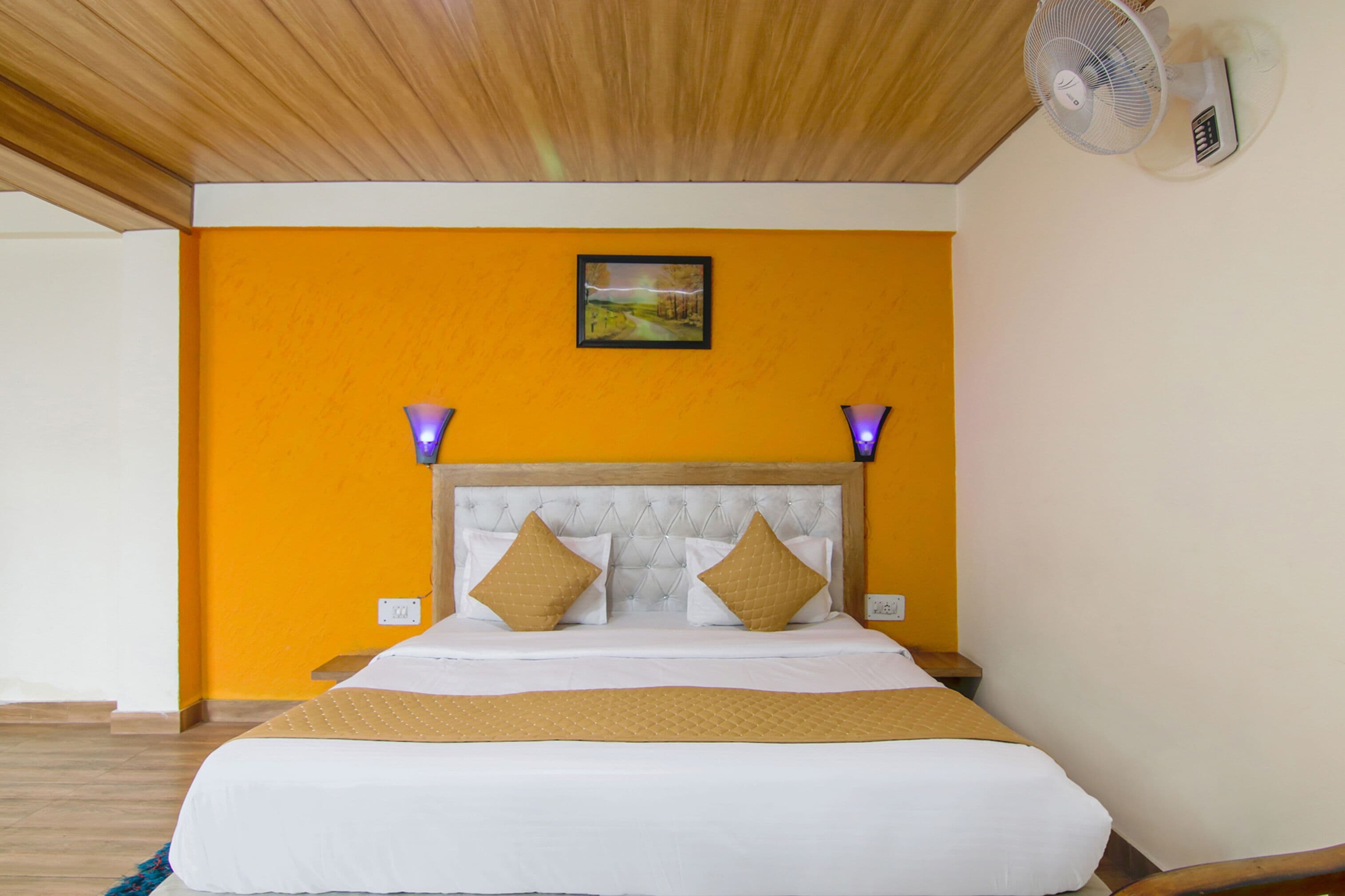 Ein Hotelzimmer mit Holzbalkendecke, einer orangefarbenen Akzentwand, einem Bett mit weißer Bettwäsche und zwei braunen Kissen sowie einem an der Wand befestigten Ventilator.