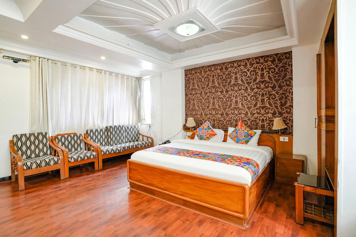 A hotel room with a wooden bed, two patterned armchairs, a large window with curtains, and a decorative headboard.