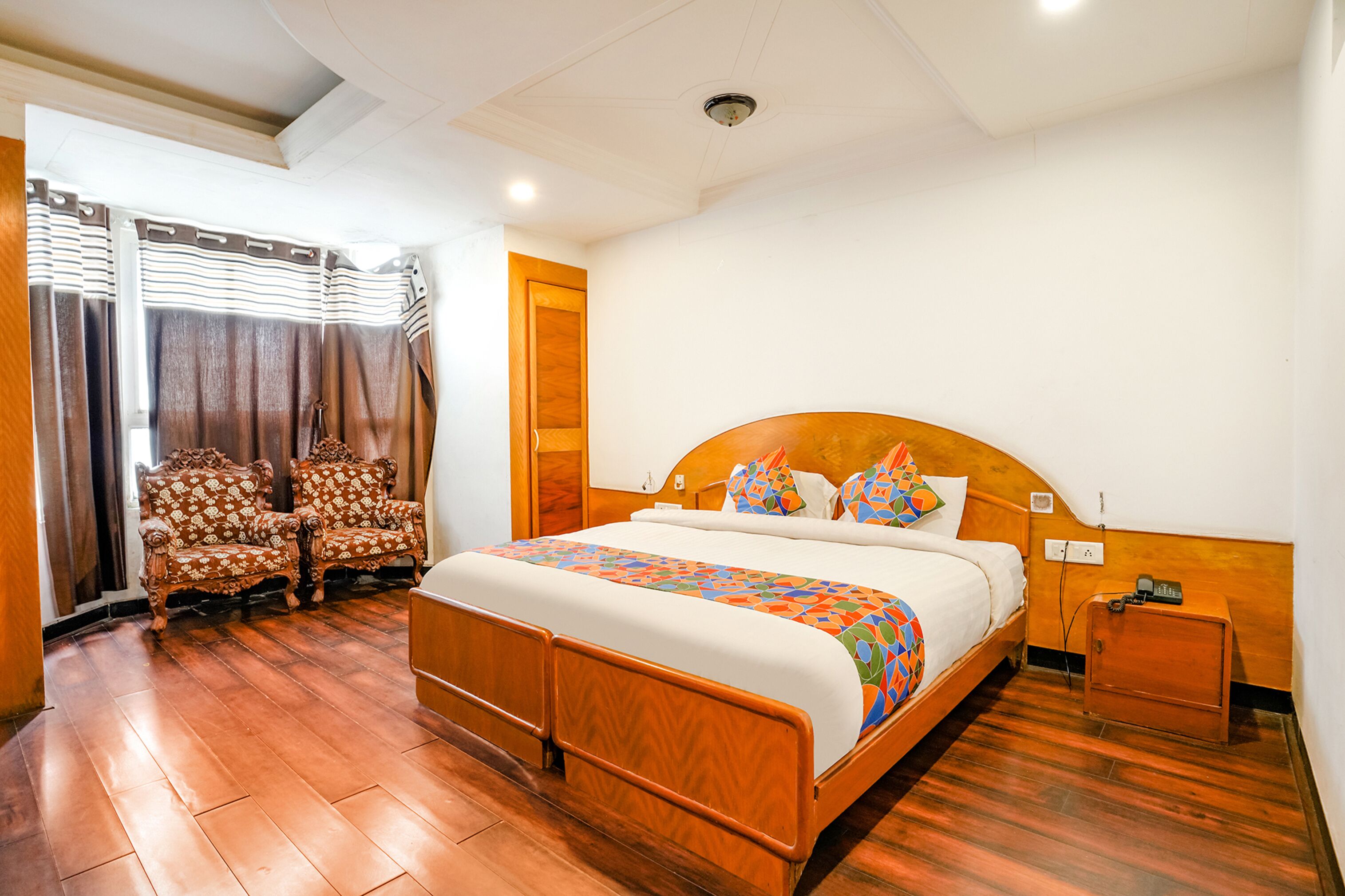 A hotel room with a wooden bed, two upholstered chairs, a nightstand, and a window with curtains.