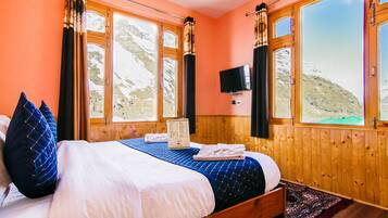 A bedroom with a wooden bed, a view of a mountainous landscape through large windows, and a flat-screen TV mounted on the wall.