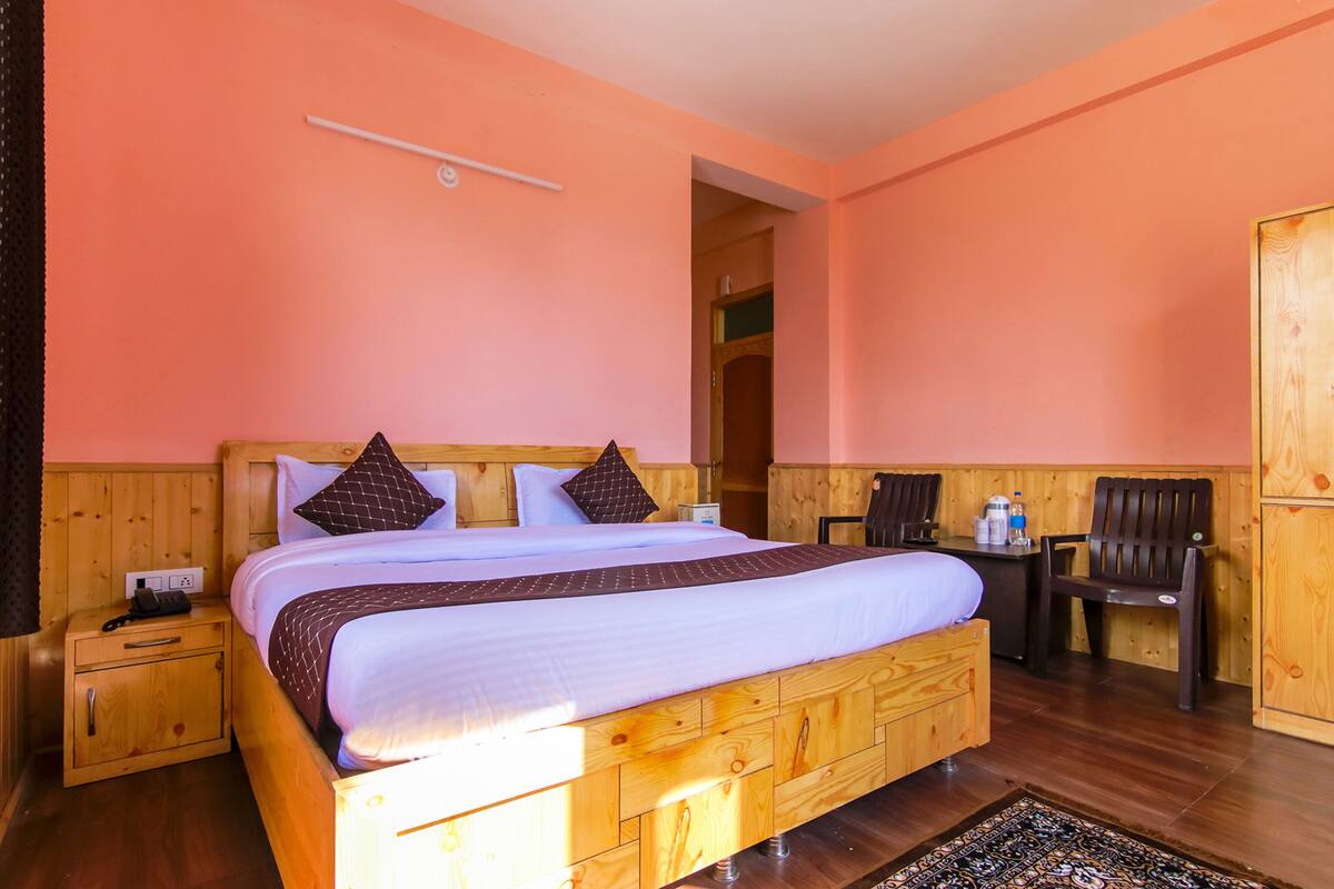 Una cama de madera con una colcha blanca y marrón, una mesita de noche con un teléfono y una silla en una habitación con paredes color melocotón y pisos de madera.