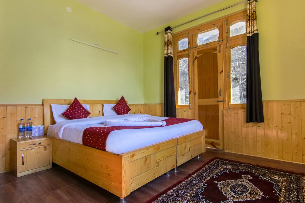 Una cama de madera con colcha roja y blanca, una mesita de noche de madera con botellas y una ventana grande con cortinas.