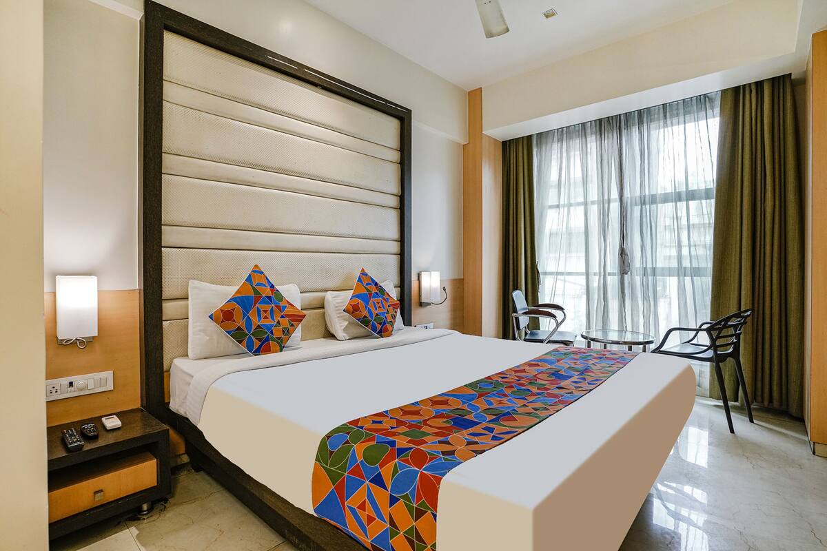 A hotel room with a large bed, a bedside table with a lamp, a chair, and a window with curtains.