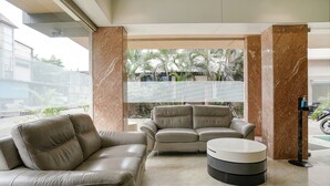 A modern living room with a beige leather sofa, a matching armchair, and a round coffee table. The room features marble columns and a large window with a view of palm trees and a building outside. - Fabhotel Konar Inn (Navi Mumbai)