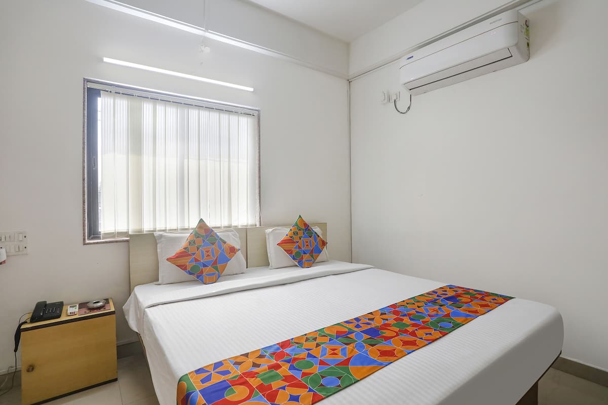A hotel room with a bed, a bedside table with a phone and a small item, a window with blinds, and an air conditioning unit on the wall.