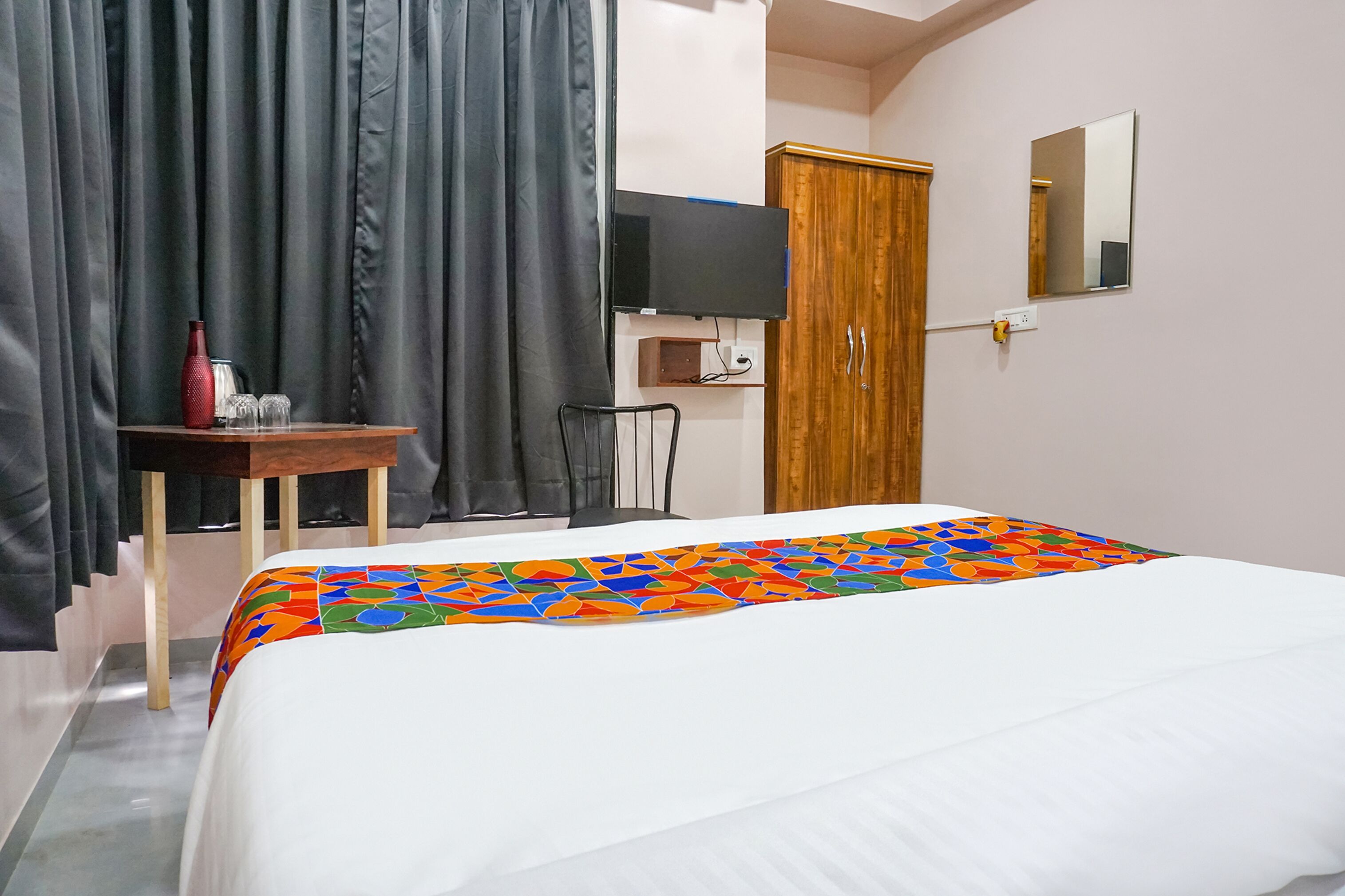 Habitación de hotel con cama, televisor, armario de madera, una mesita con un florero rojo y un espejo en la pared.