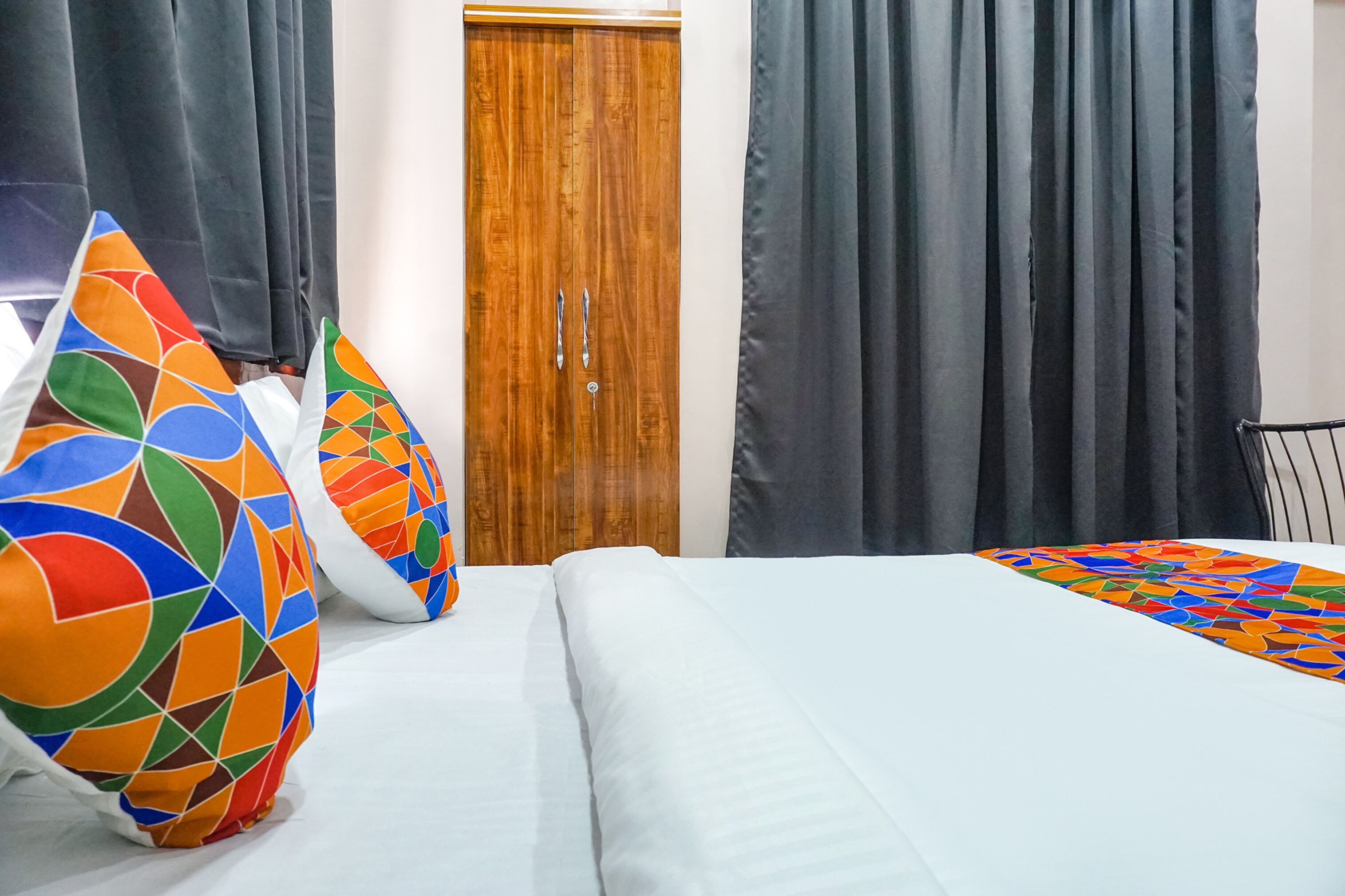 A hotel room with a wooden wardrobe, a bed with white bedding, and colorful geometric-patterned pillows.