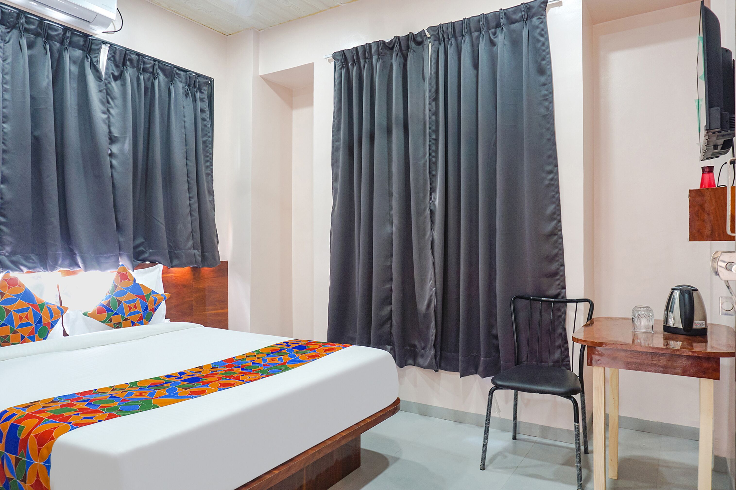 A hotel room with a bed, a chair, a table with a kettle, and curtains.