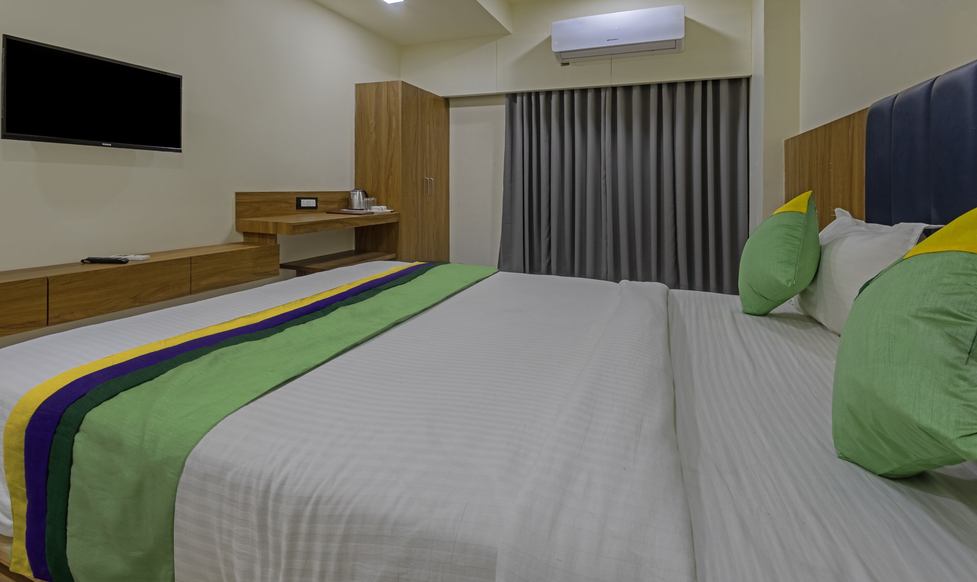 A hotel room with a large bed, a wooden nightstand, a flat-screen TV mounted on the wall, and a window with curtains.