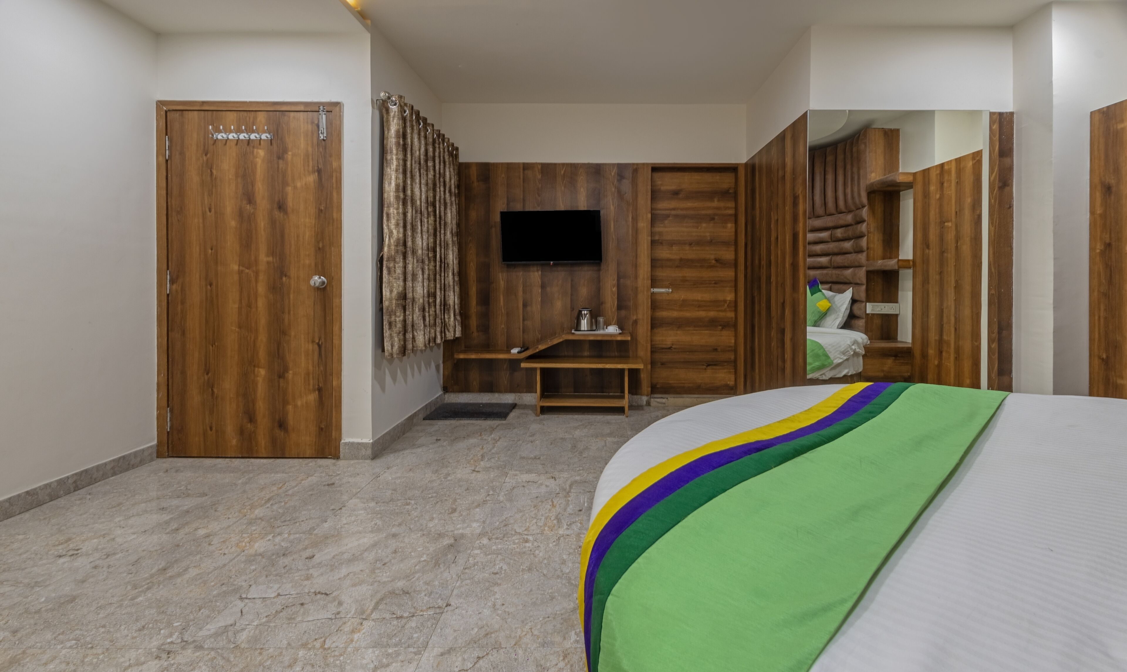 A hotel room with a wooden door, a television mounted on the wall, a bed with a colorful blanket, a small wooden bench, and a bedside table with a kettle and cups.