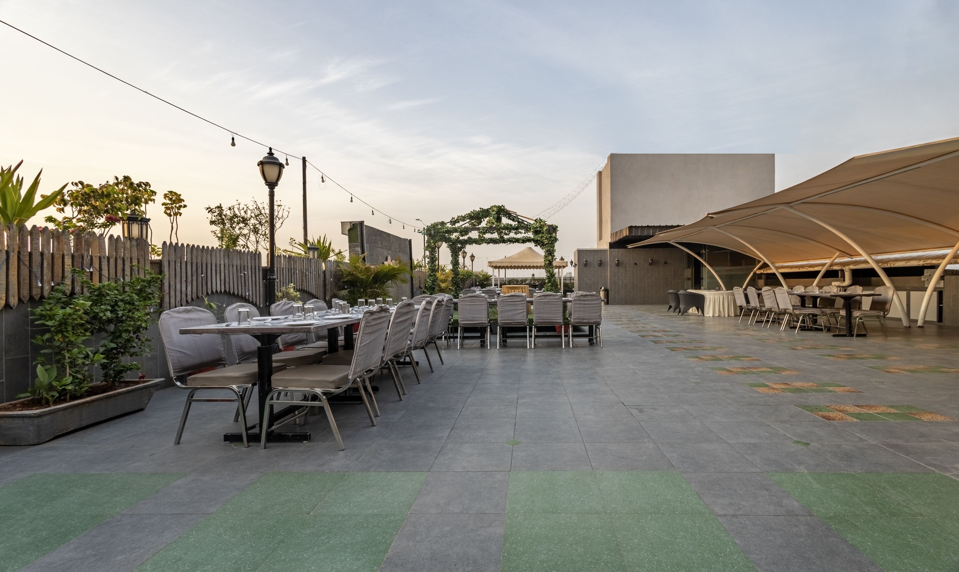 An outdoor dining area with tables and chairs, a pergola with greenery, and a modern building in the background.
