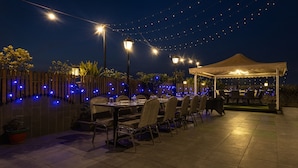 Een terras met tafels en stoelen, sfeerverlichting en een zonnescherm.