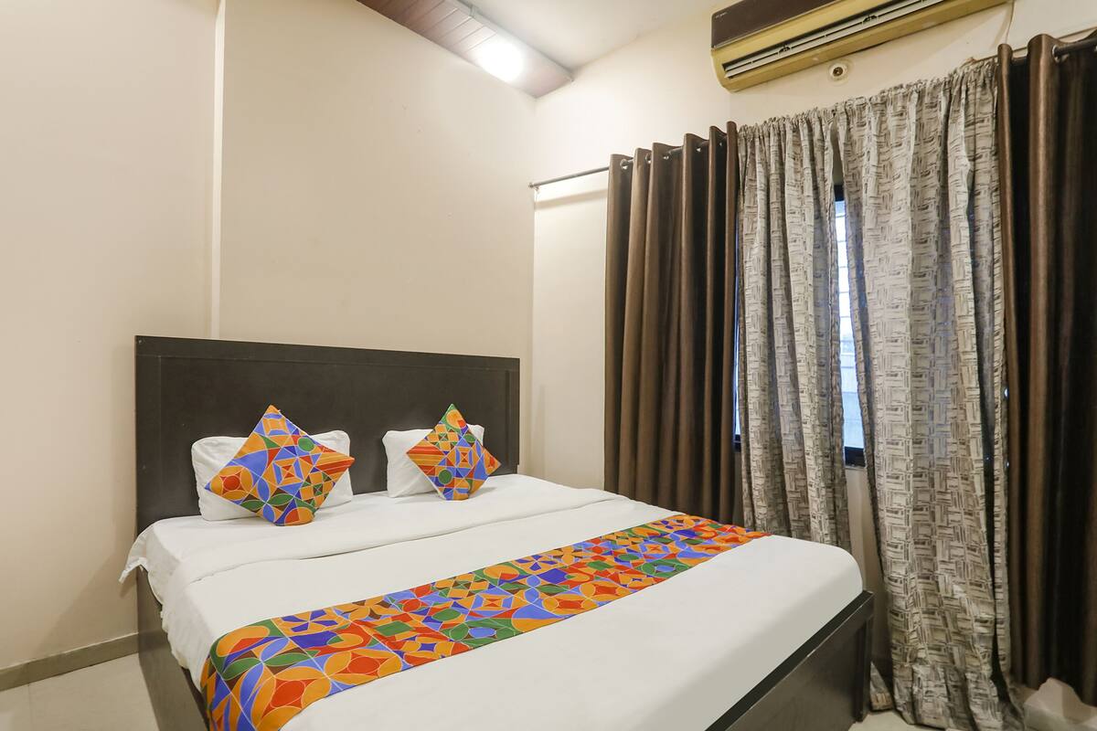 Una habitación de hotel con una cama, dos almohadas, una colcha colorida, cabecera, una ventana con cortinas y un equipo de aire acondicionado.