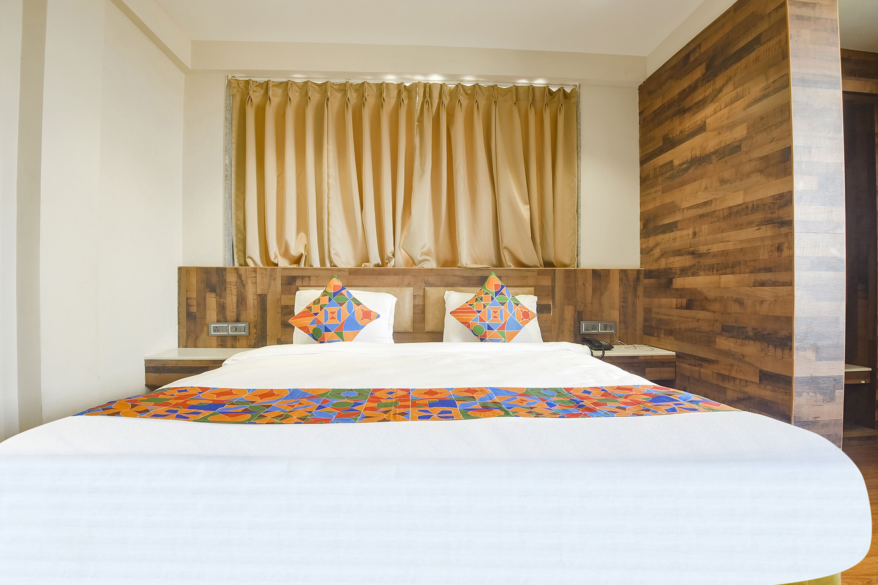A hotel room with a large bed, a wooden headboard, a window with curtains, and a nightstand with a lamp.