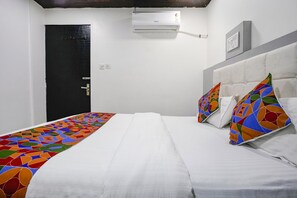 A hotel room with a bed, white bedding, colorful pillows, a black door, and an air conditioning unit on the wall.