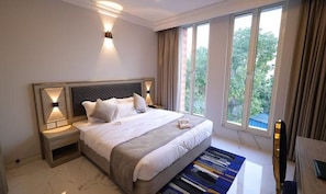 Ein modernes Hotelzimmer mit einem großen Bett, Nachttischlampen, Blick ins Grüne durch die Fenster und einem kleinen Schreibtisch mit Stuhl.