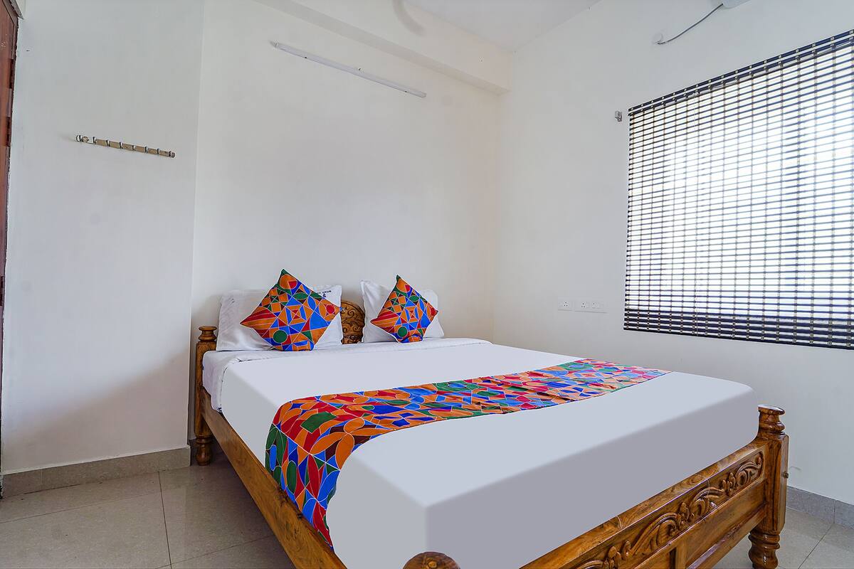 A neatly made bed with a colorful blanket and patterned pillows, a wooden headboard, and a window with blinds.