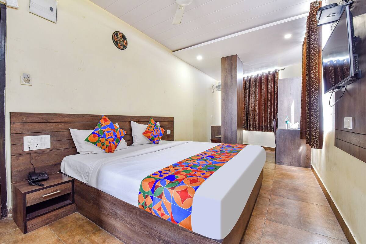 Una habitación de hotel con cabecera de madera, cama con ropa de cama geométrica de colores, mesita de noche con teléfono, baño con cortina de ducha y televisor montado en la pared.