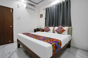 A neatly made bed with a colorful geometric-patterned bedspread, a wooden headboard, a nightstand with a lamp, and a window with dark curtains. - Fabhotel Fortuna Inn (Navi Mumbai)