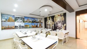 A dining area with white tables and chairs, a large wall mural of temples, and a ceiling with recessed lighting. - FabHotel Aditya Homes (Hyderabad)