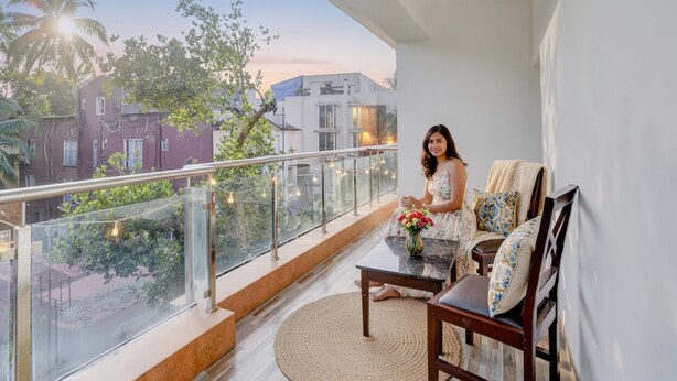A balcony with a glass railing, a wooden chair, a small table with a vase of flowers, and a view of buildings and trees. - FabHotel The Sky - Nr. Calangute Beach (Calangute)