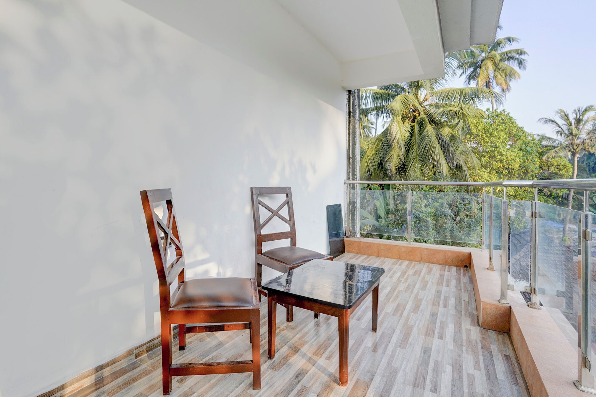 A balcony with two wooden chairs and a small table, overlooking a tropical landscape with palm trees.