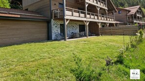 Un chalet en bois à deux étages, doté d’un balcon, de fondations en pierre et d’une cour gazonnée.
