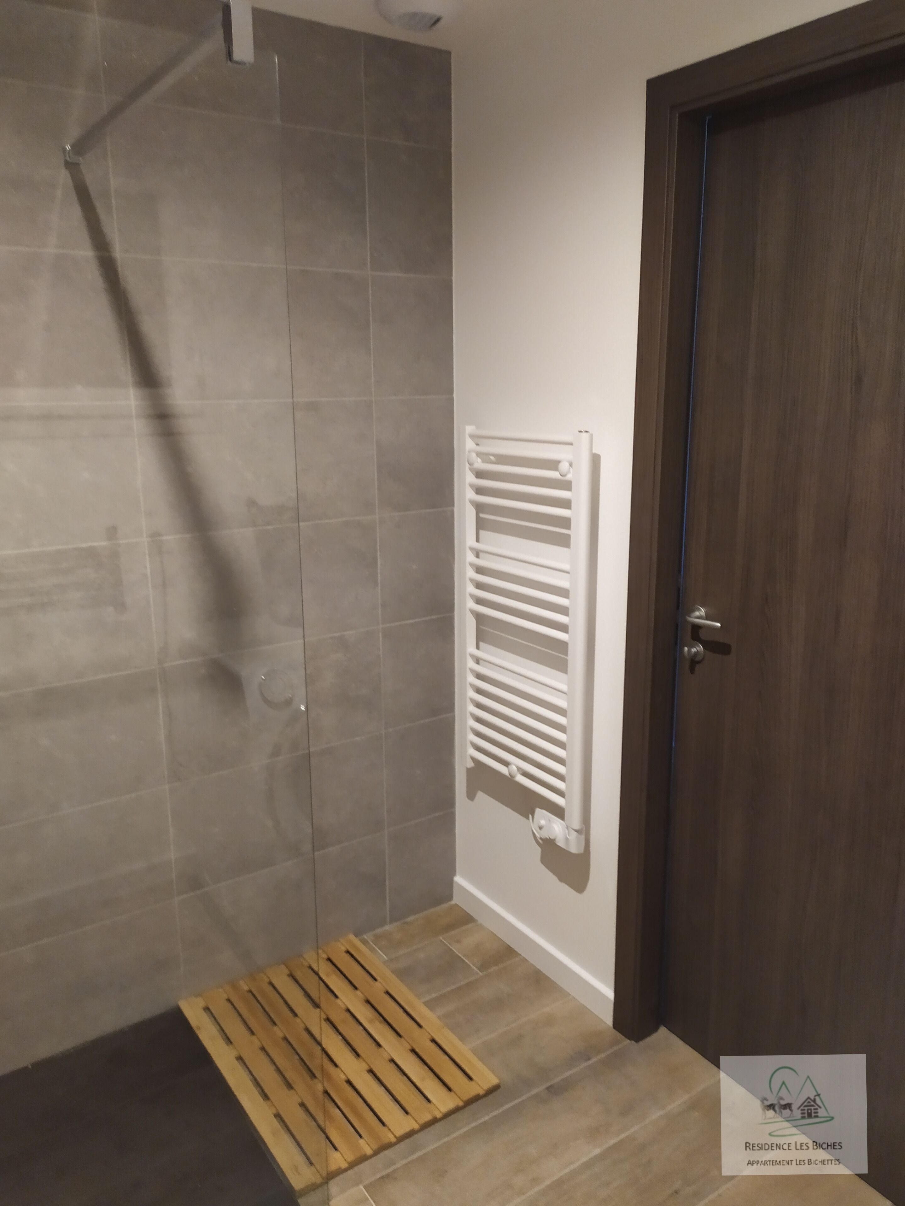 Un área de ducha con pared de azulejos, suelo de madera, un toallero blanco y una puerta de madera.