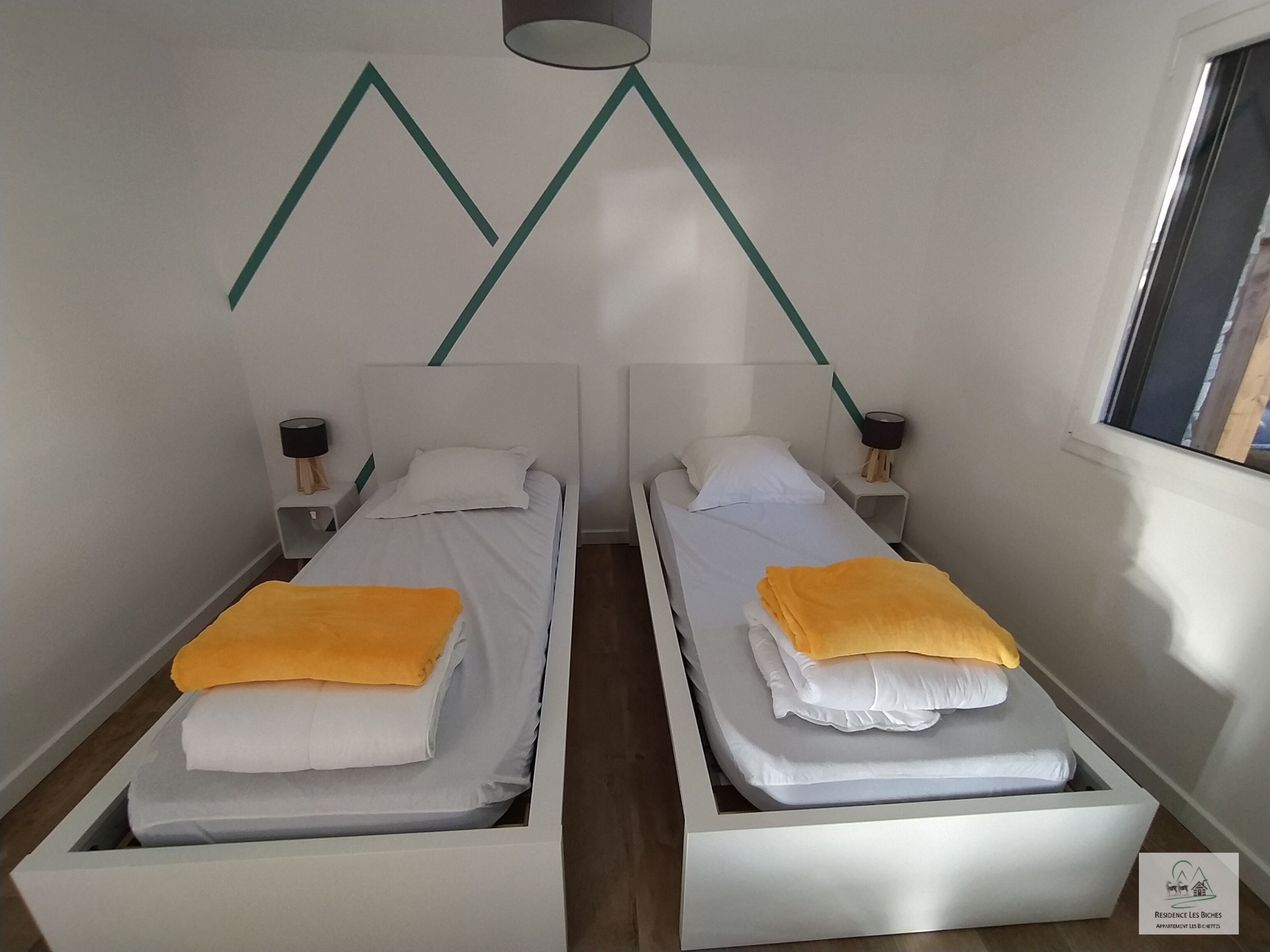 A hotel room with two single beds, each with a white mattress and a yellow blanket. There are two bedside tables with lamps, and the walls are painted white with a green geometric design.