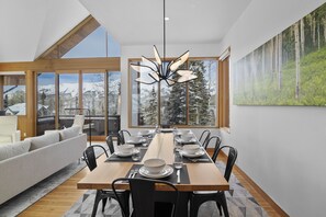 A dining area with a long wooden table set for six, surrounded by black chairs. Large windows offer a view of snow-capped mountains and a forested area.
