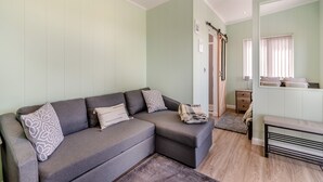 Un salon comprenant un canapé d’angle, une petite table et un banc. Une porte donne accès à une chambre.