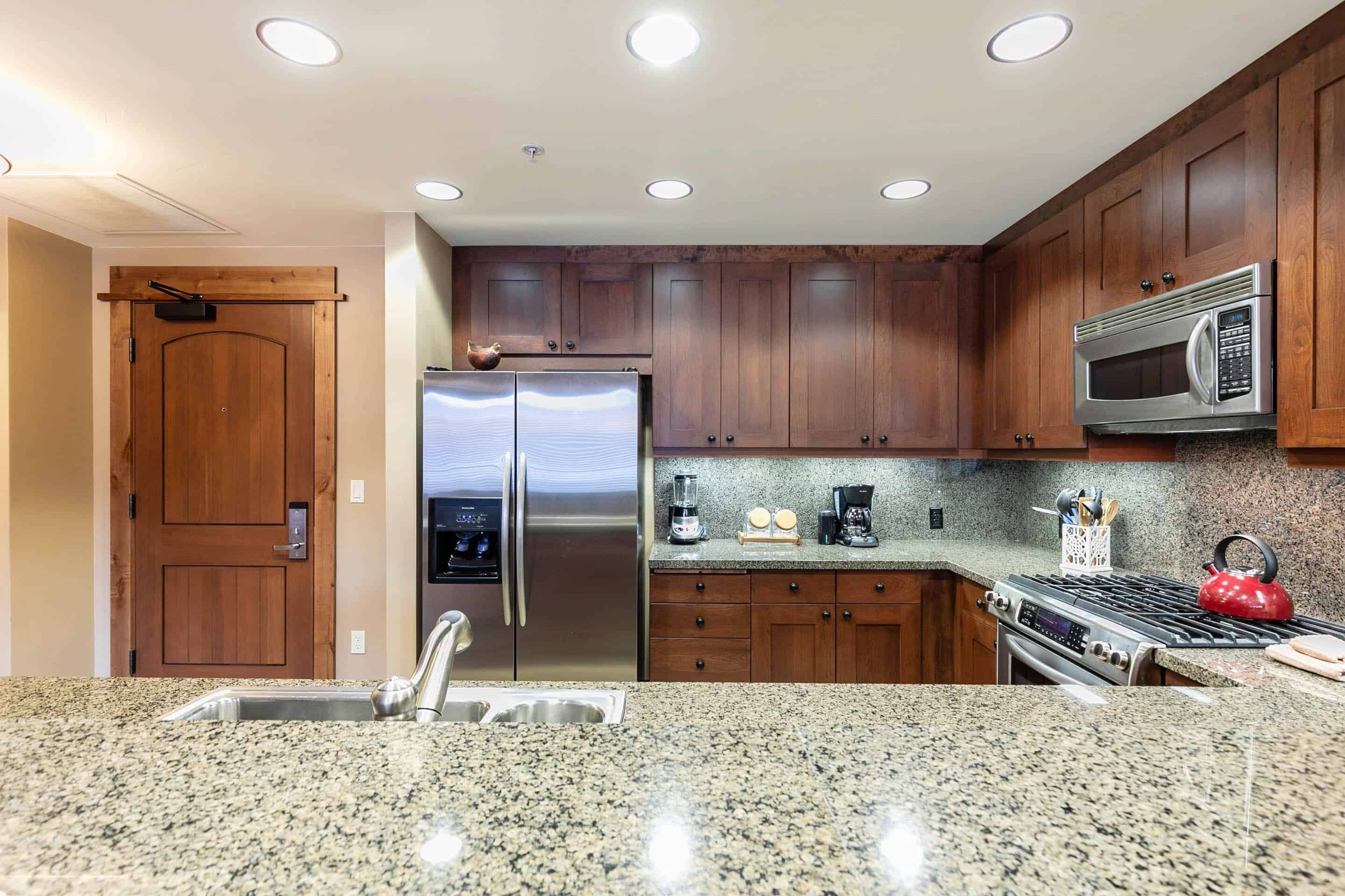 Una cocina con encimeras de granito, electrodomésticos de acero inoxidable, armarios de madera y una tetera roja.