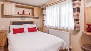 A bedroom with a bed, a radiator, a window with curtains, a shelf with items, and a chair.