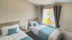 Habitación doble con dos camas, ventana con cortinas, mesita de noche con lámpara y radiador.