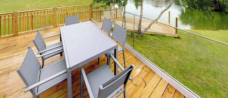 A deck with a table and chairs, overlooking a body of water and a grassy area with trees.