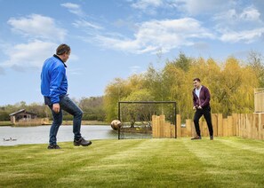 Deux personnes jouent au football sur un terrain herbeux, avec des buts de football, une étendue d’eau et des arbres en arrière-plan.