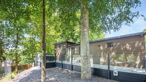 A modern RV with a covered deck, surrounded by trees and gravel. - The Watchtower (Pet) (Snowdonia)