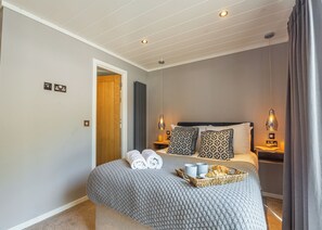 A hotel room with a large bed, two bedside lamps, a bedside table with a tray of croissants, and a door leading to another room.
