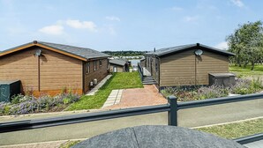 Une rangée de cabanes séparées par un chemin pavé, une zone herbeuse parsemée de fleurs et un ciel dégagé.