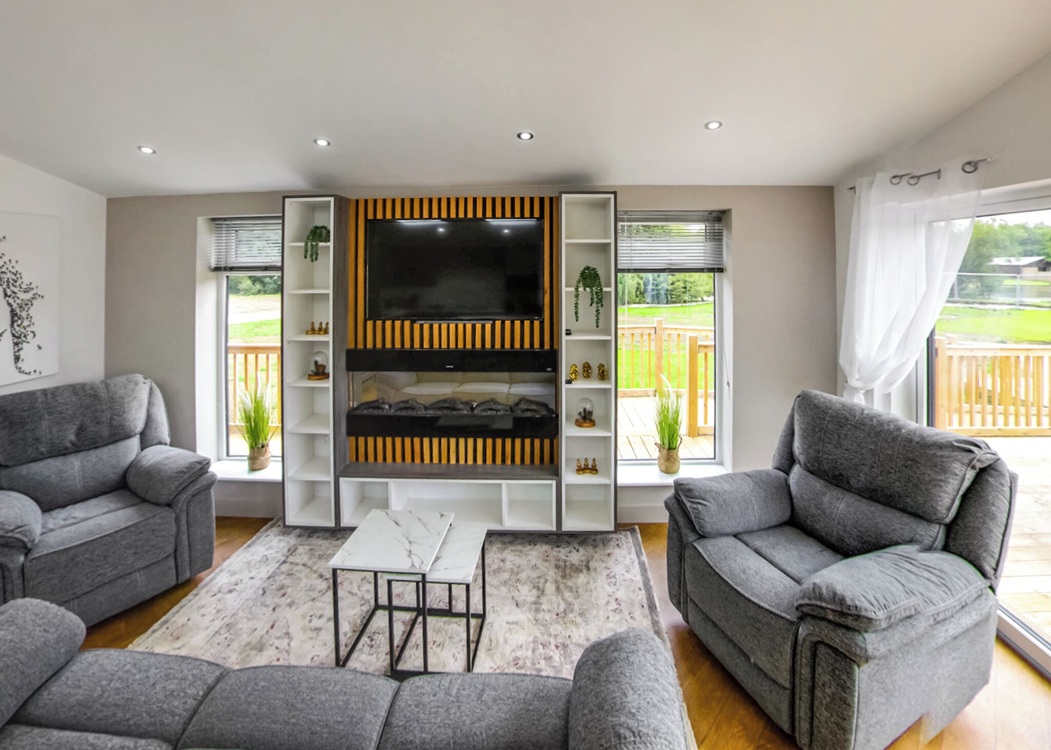Een woonkamer met grijze banken, een flatscreen-televisie, planken en uitzicht op een houten terras.