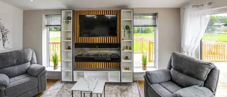 A living room with grey sofas, a flat-screen TV, shelves, and a view of a wooden deck.