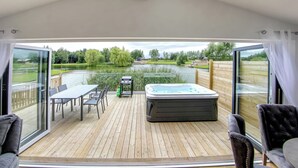 A deck with a hot tub, table, chairs, and a view of a lake and greenery. - Superb bespoke lodge with two bedrooms sleeping up to four people. (Laceby)