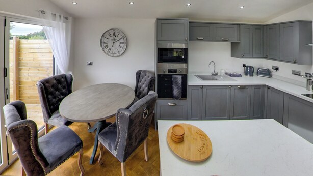 A modern kitchen with grey cabinets, a round dining table, and a clock on the wall. - Superb bespoke lodge with two bedrooms sleeping up to four people. (Laceby)