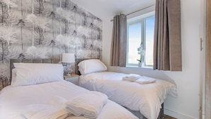 A hotel room with two beds, a nightstand with a lamp, a window with curtains, and a wall with a tree pattern. - Superb bespoke lodge with two bedrooms sleeping up to four people. (Laceby)